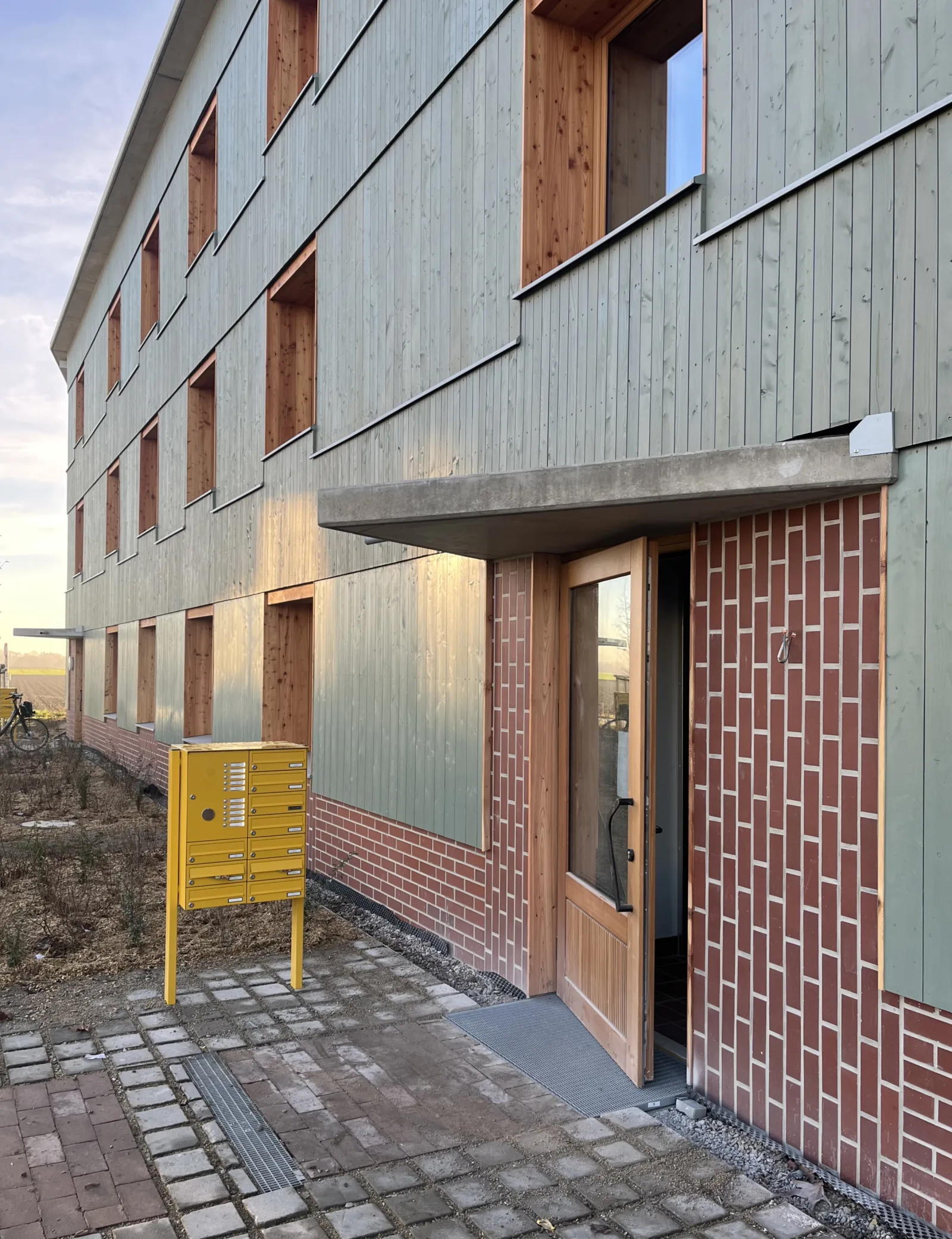 Hauseingang mit Holzfassade, Ziegelsockel und gelber Briefkastenanlage; davor ein Weg aus unterschiedlich großen Pflastersteinen.