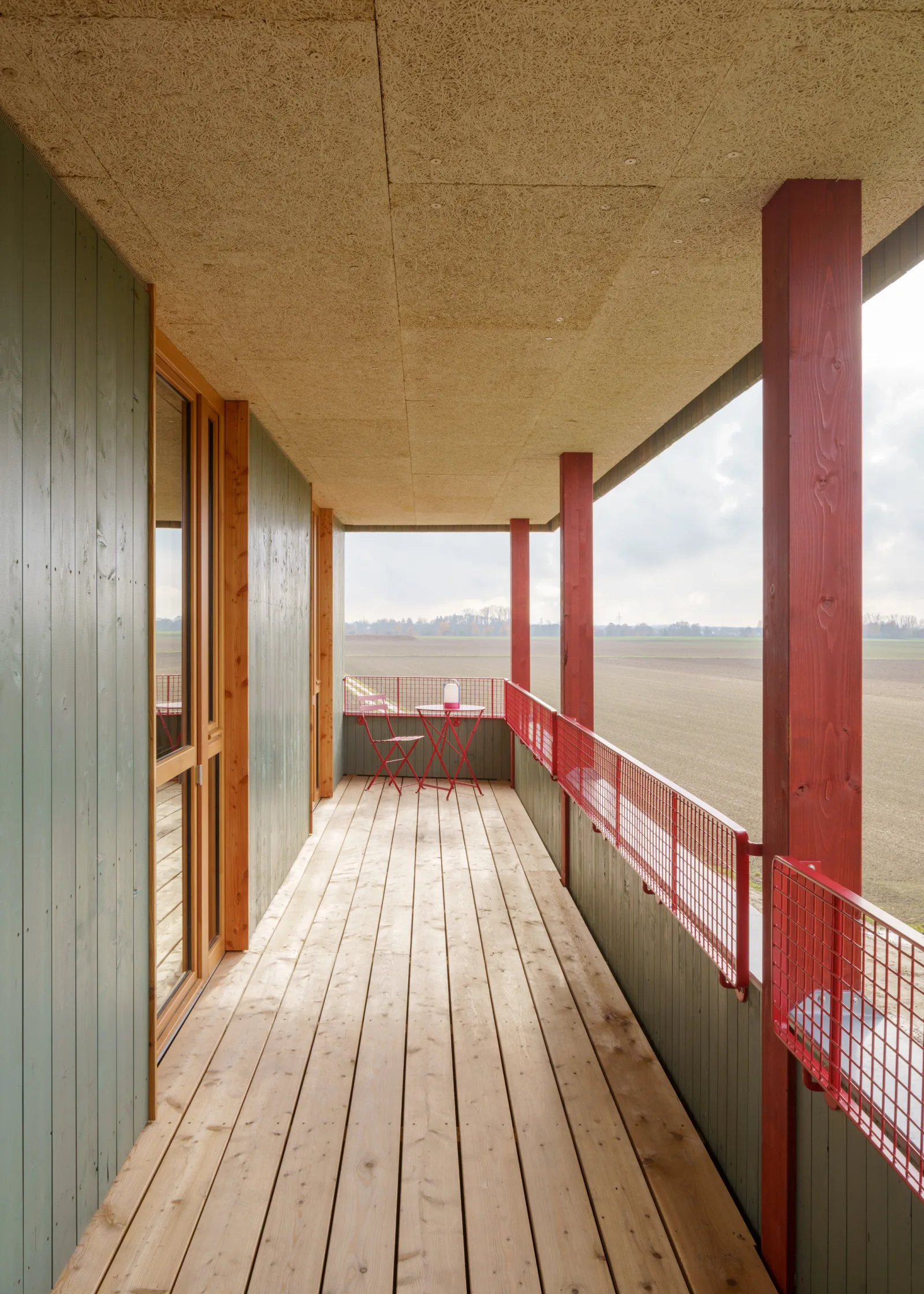 Überdachter Balkon mit Holzdielen, roten Holzstützen und Metallgeländer; die Deckenplatten sind sichtbar verschraubt.