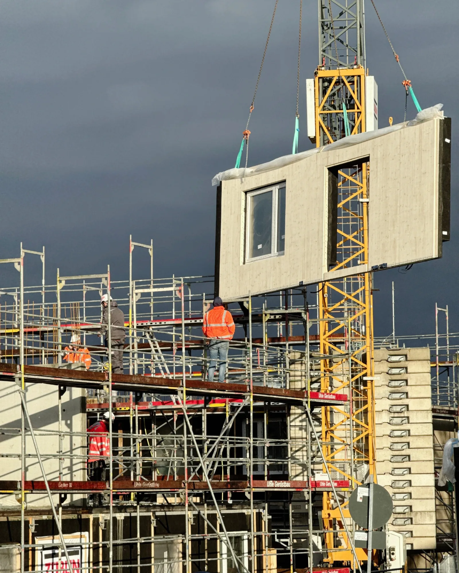 Ein vorgefertigtes Wandelement mit Fensteröffnung hängt am Kran vor dem eingerüsteten Rohbau